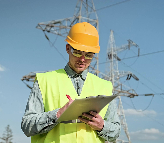 A man in a green vest and hard hat holds a clipboard, standing in a construction site