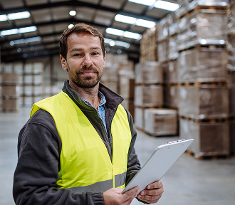 A man in a green vest holds a clipboard, standing and preparing for a task in a warehouse
